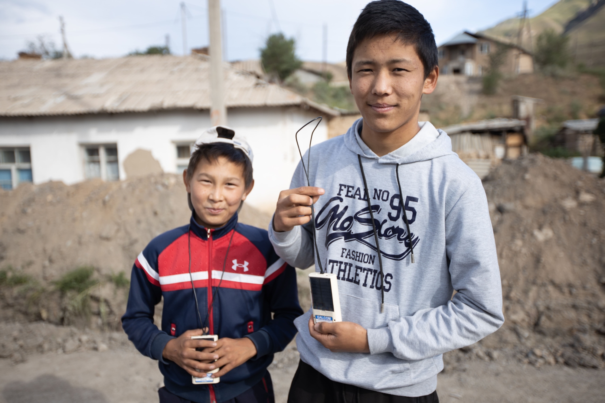 Two smiling teenage boys holding portable audio devices in Kyrgyzstan.