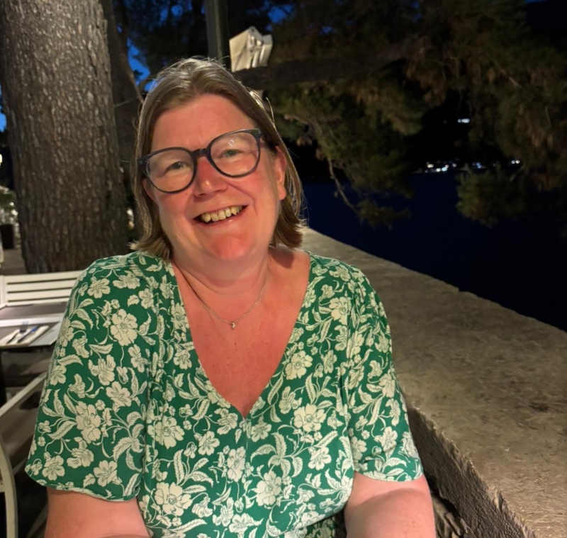 Catherine sitting outside on a terrace at night, smiling. She has a brown bob and is wearing black rimmed glasses and a green floral dress.