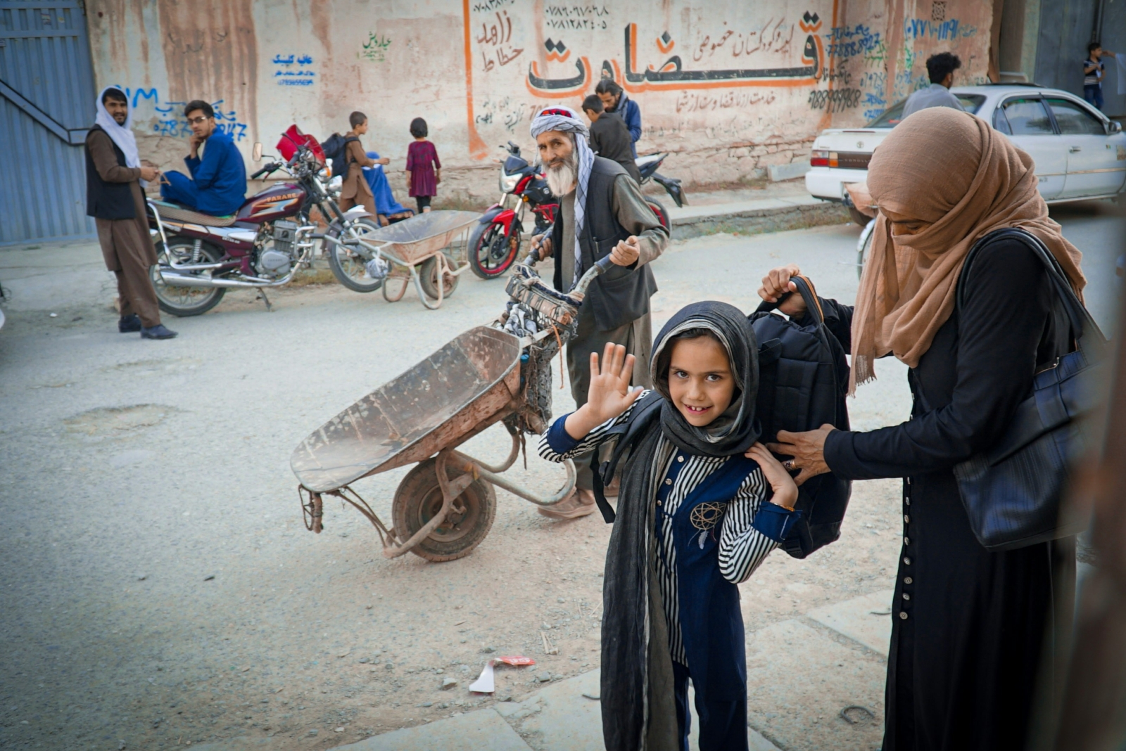A young Afghan girl waves at the camera as her mum helps her put on a black backpack on a busy street.