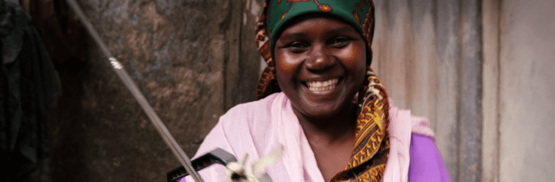 A woman in Mozambique grins at the camera while holding a radio. She is wearing a paisley green scarf on her head and a pink and purple top.