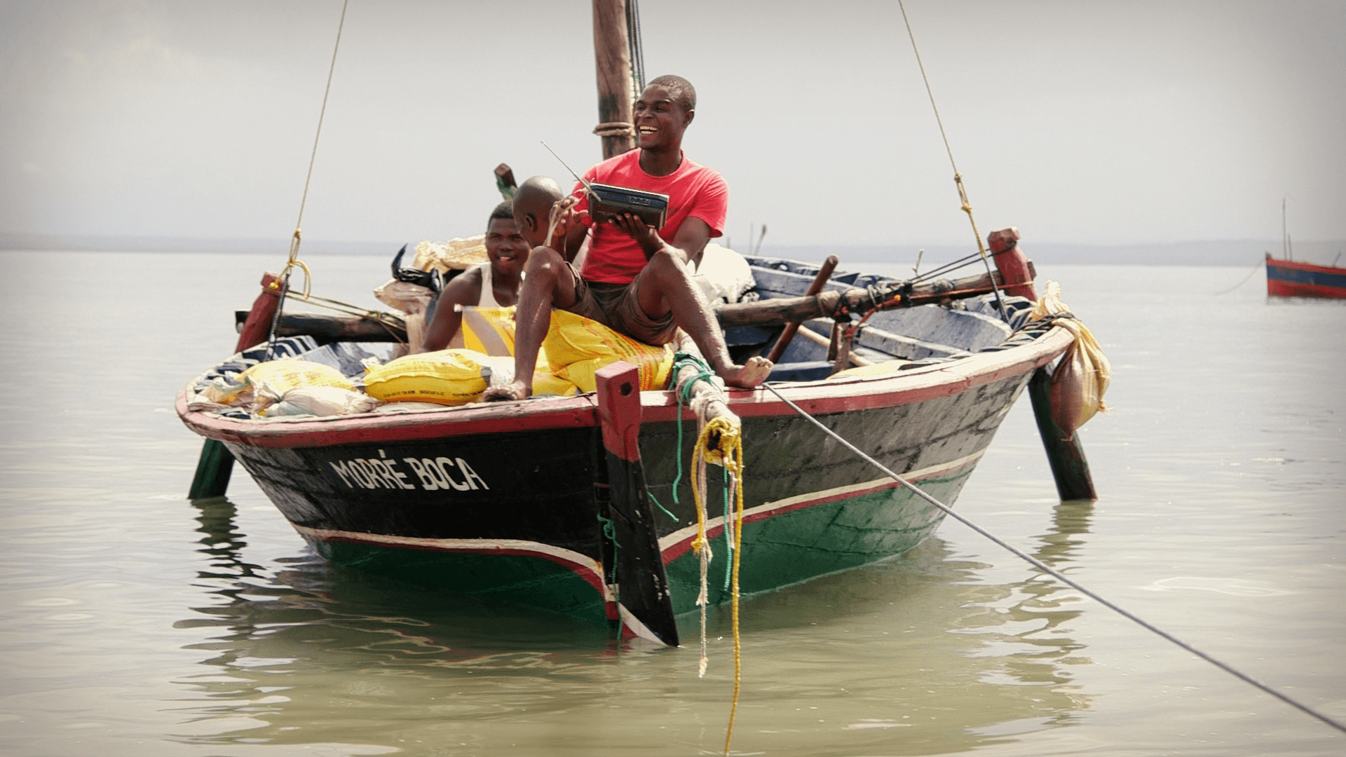 A young man sits on the front a small fishing boat floating on the water laughing as he listens to a radio.