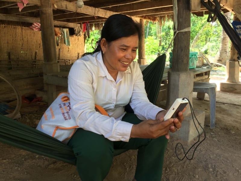 A female listener sitting on a hammock in a rural village in Cambodia. She is smiling as she holds and listens to a small white audio device.