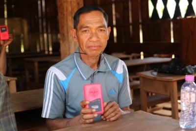 A portrait of man sitting in a wooden building in Cambodia holding a red audio Bible.