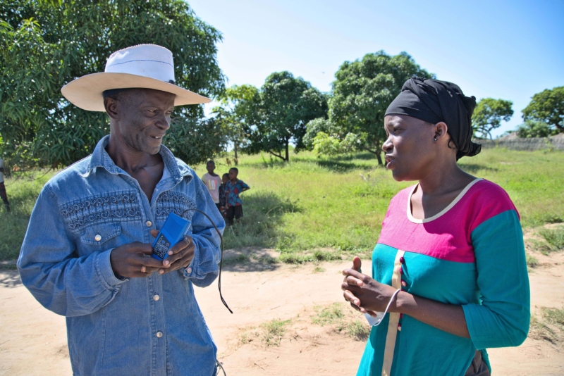 Farida, Station Manager at Radio Wimbe in Mozambique, talking to a man holding a small blue audio device. They are standing outside surrounded by grass and trees.