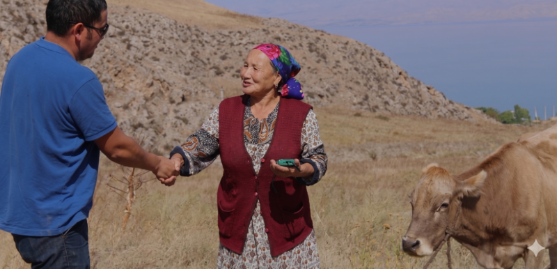 An older woman shaking hands with a man in a blue top who has given her a small audio device. They are standing in a field on the side of a hill in Kyrgyzstan with a cow behind them.
