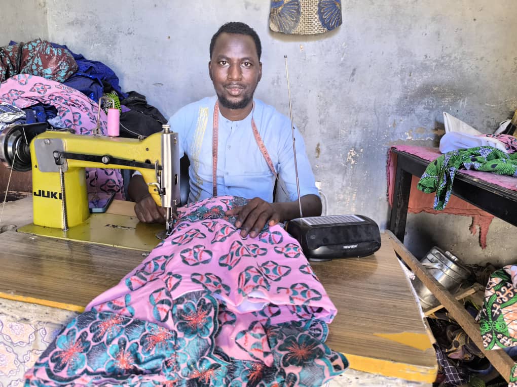A young man sewing at an old fashioned yellow sewing machine while listening to a solar radio in Mali.
