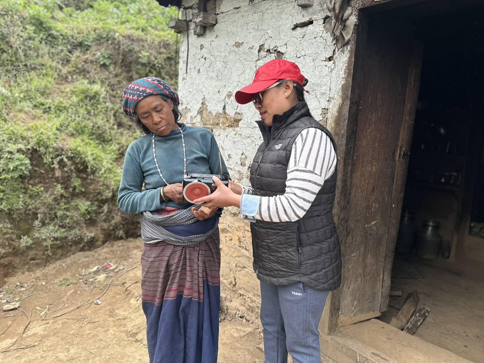Binu, a member of the Afno FM team in Nepal, shows a female listener how to use a small black radio outside a simple, white house.