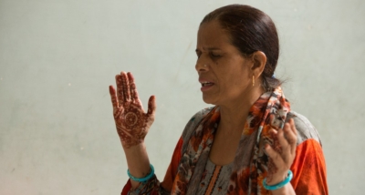 An Indian woman stand with her eyes closed and hands lifted in passionate prayer. She has henna patterns on her hands and is wearing a matching top and scarf around her neck.