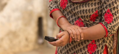 Close up shot of an Indian woman's hands holding a portable life changer speaker box. She is wearing a brown and red embroidered dress with matching red bangles.