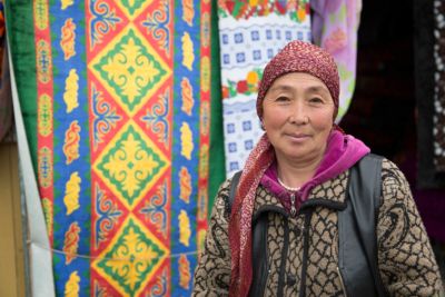 A woman wearing a red headscarf smiles with bright, colourful patterned matts hanging behind her in Kyrgyzstan.