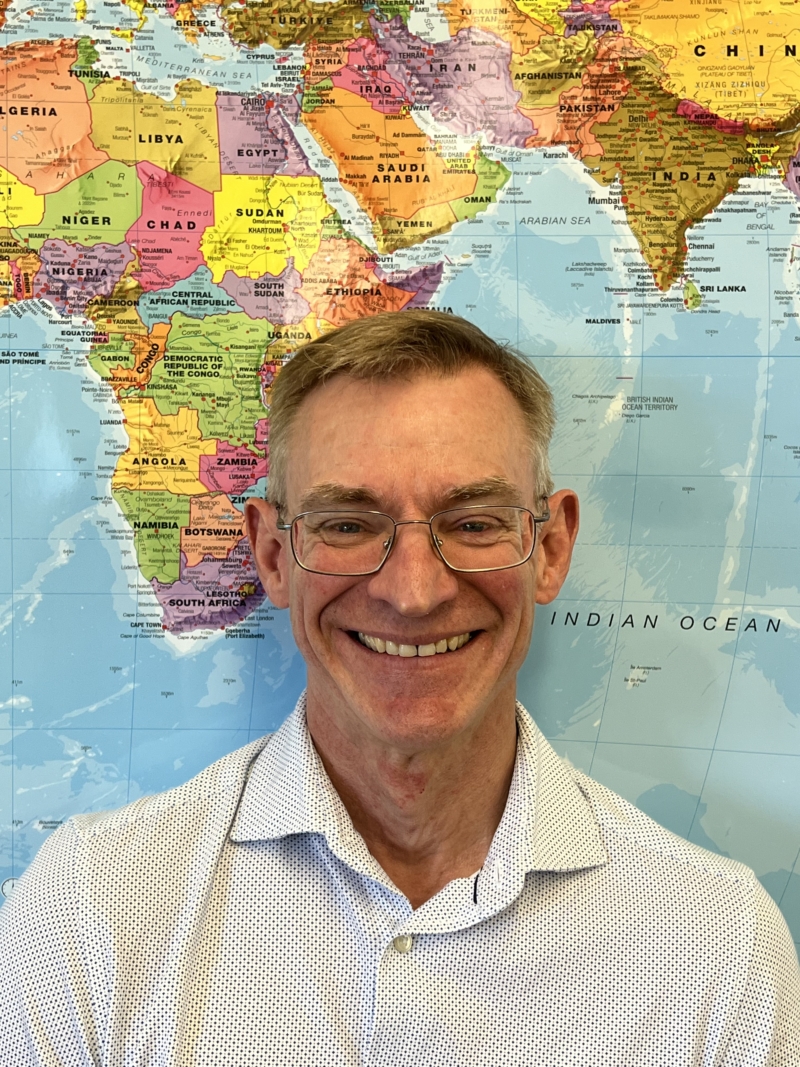 Kevin standing in front of a map of the world smiling. He has short grey hair and silver glasses and is wearing a white shirt.