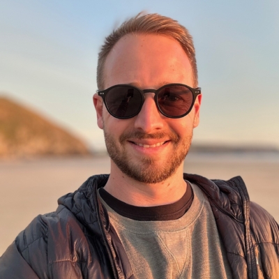 Luke smiling on a beach, wearing a pair of black sunglasses.
