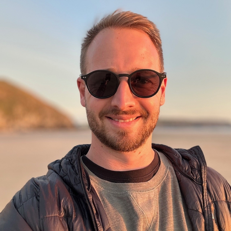 Luke smiling on a beach, wearing a pair of black sunglasses.
