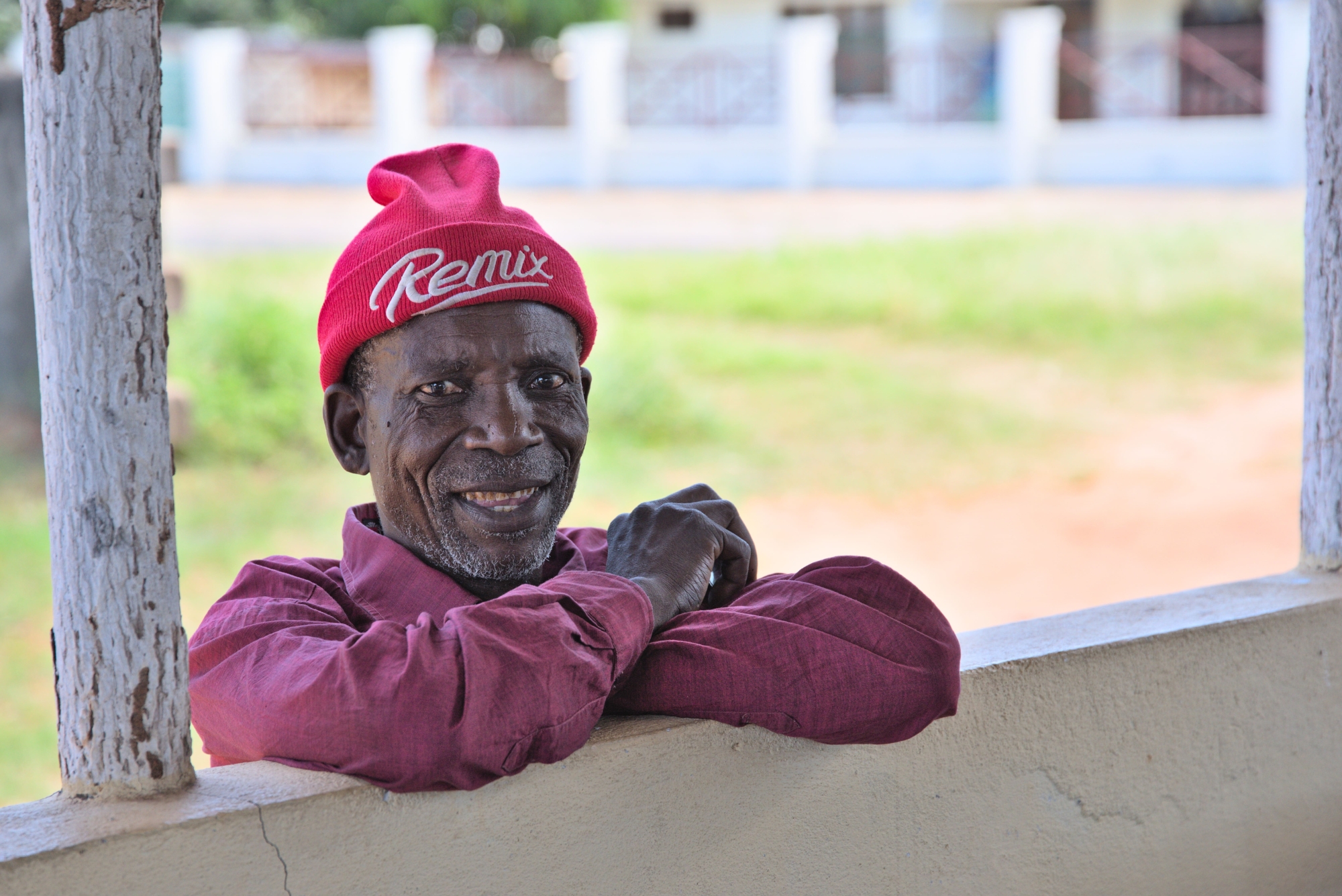 A man leaning on a wall in Mozambique. He is smiling and wearing a red shirt, and red beanie hat.