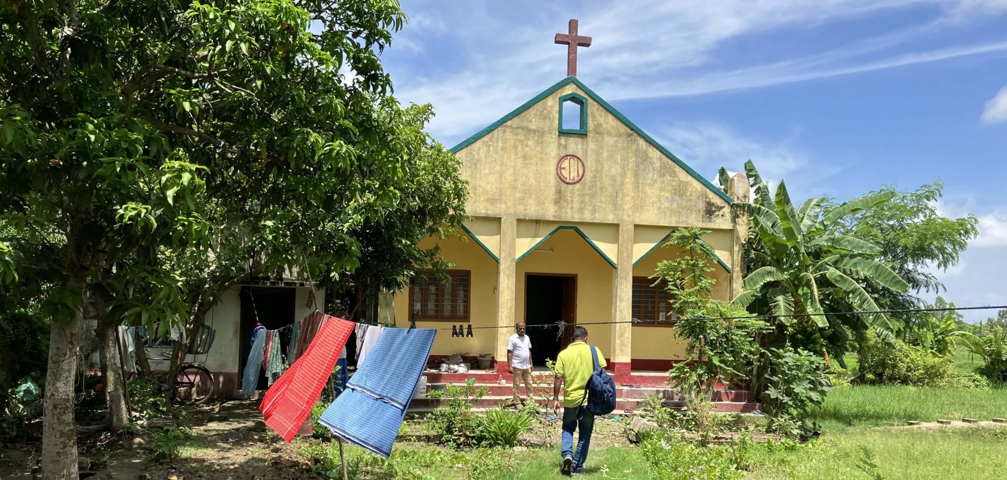 A yellow church building with a cross on top in India, surrounded by green palm trees.
