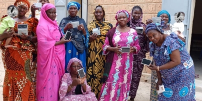 A group of women in Mali stand together holding portable solar powered audio devices. They are wearing colourful traditional dresses and matching head scarves.