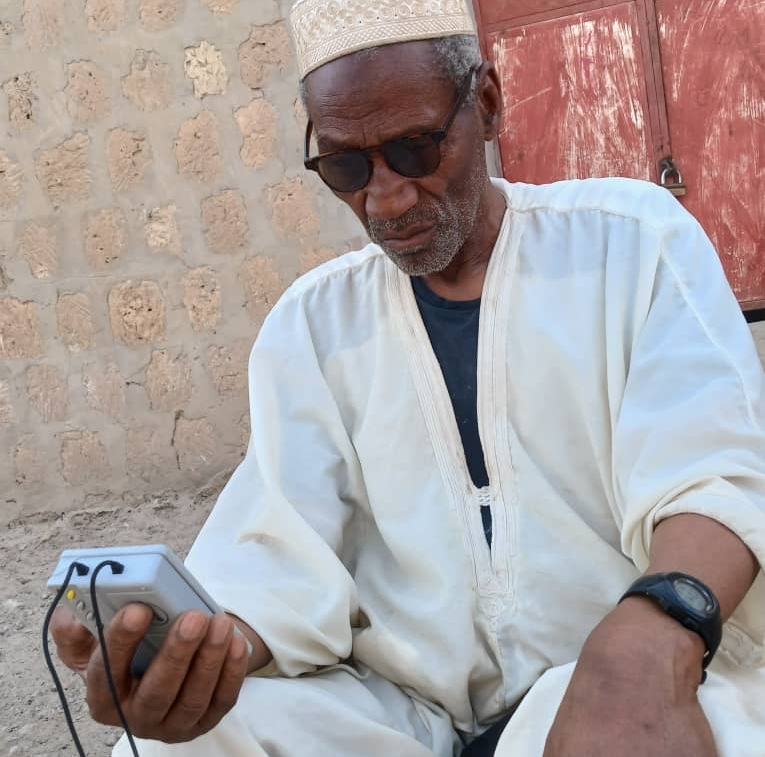 A man sits outside a stone building in Mali listening to a small audio device. He is wearing sunglasses, a traditional hat and white tunic.