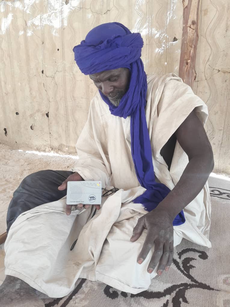 A man in a blue turban and cream tunic sits on the floor listening to a small audio device in Mali.