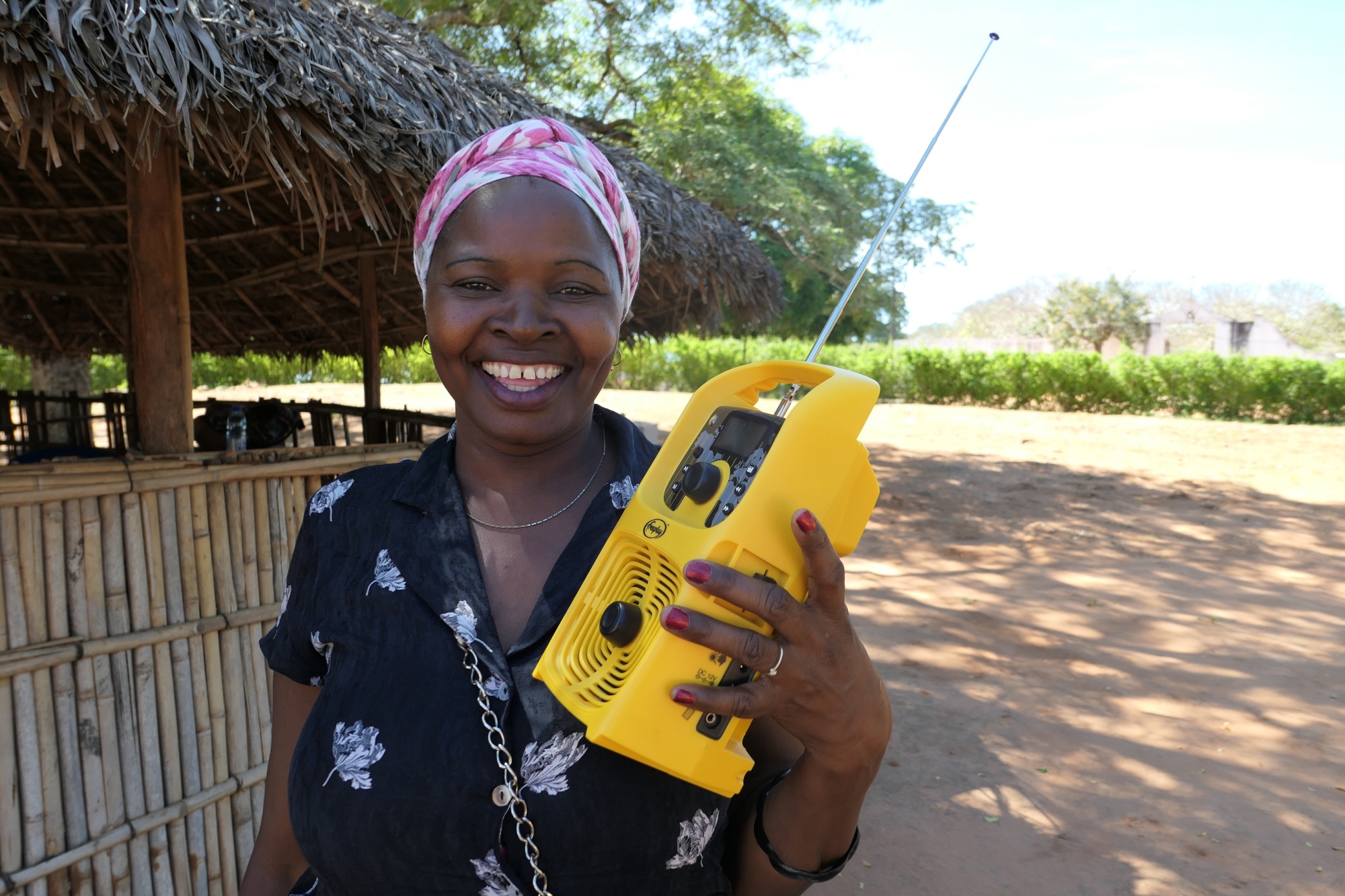 A woman smiling holding a bright yellow radio in a rural village in Mozambique. She is wearing a pink headscarf and a black shirt with flowers on it.