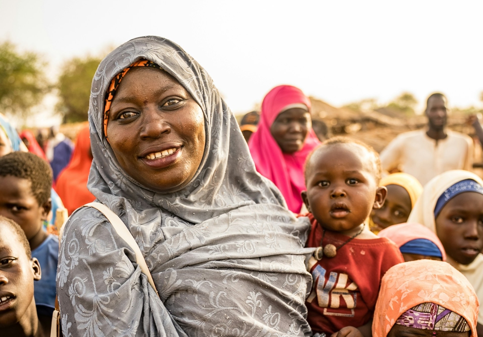 A woman in West Africa wearing a grey floral hijab holding a small baby smiles, surrounded by other people blurred in the background.