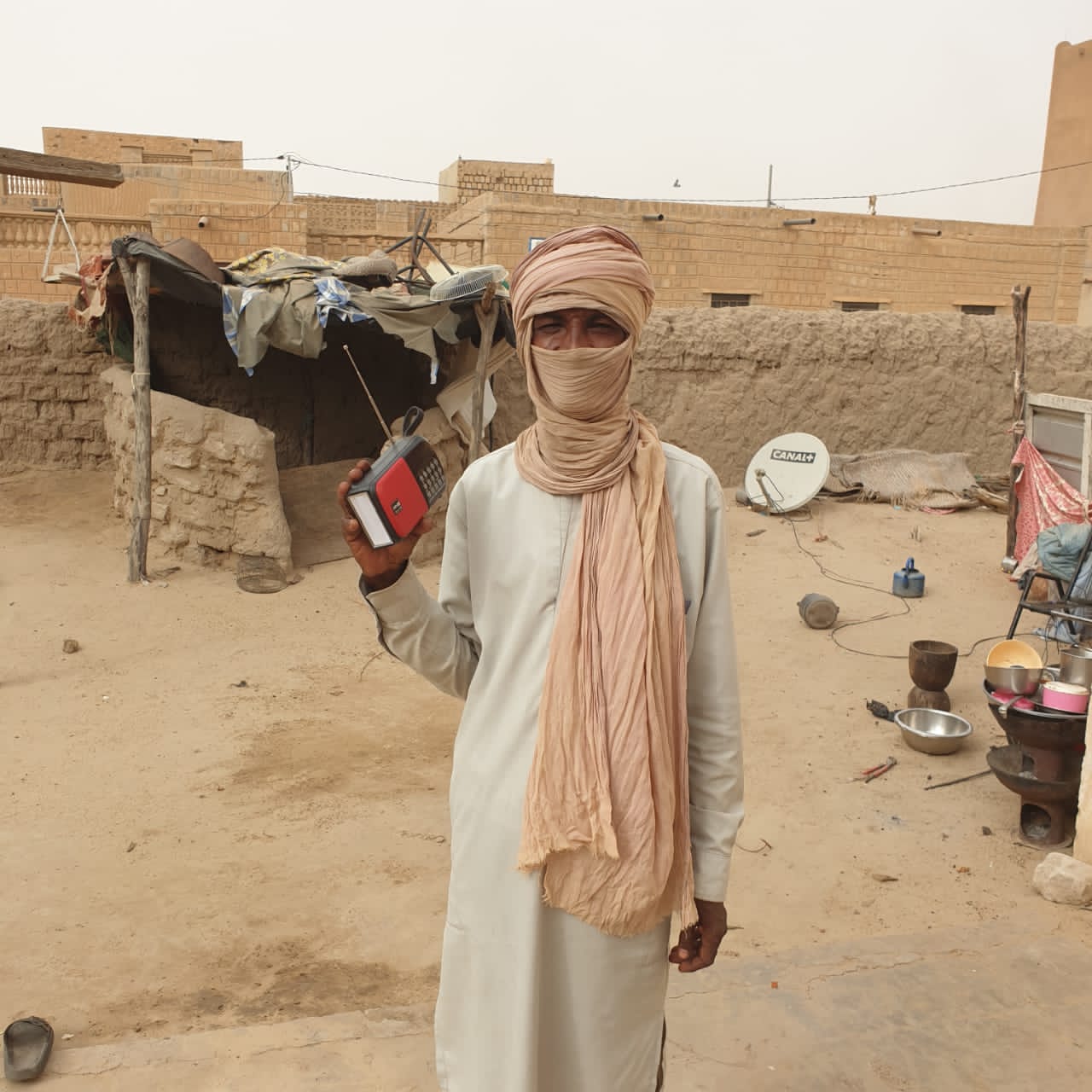 A man stand in a sandy courtyard in Mali holding a small red and black radio. He is wearing a beige turban that covers most of his face, and a white tunic.
