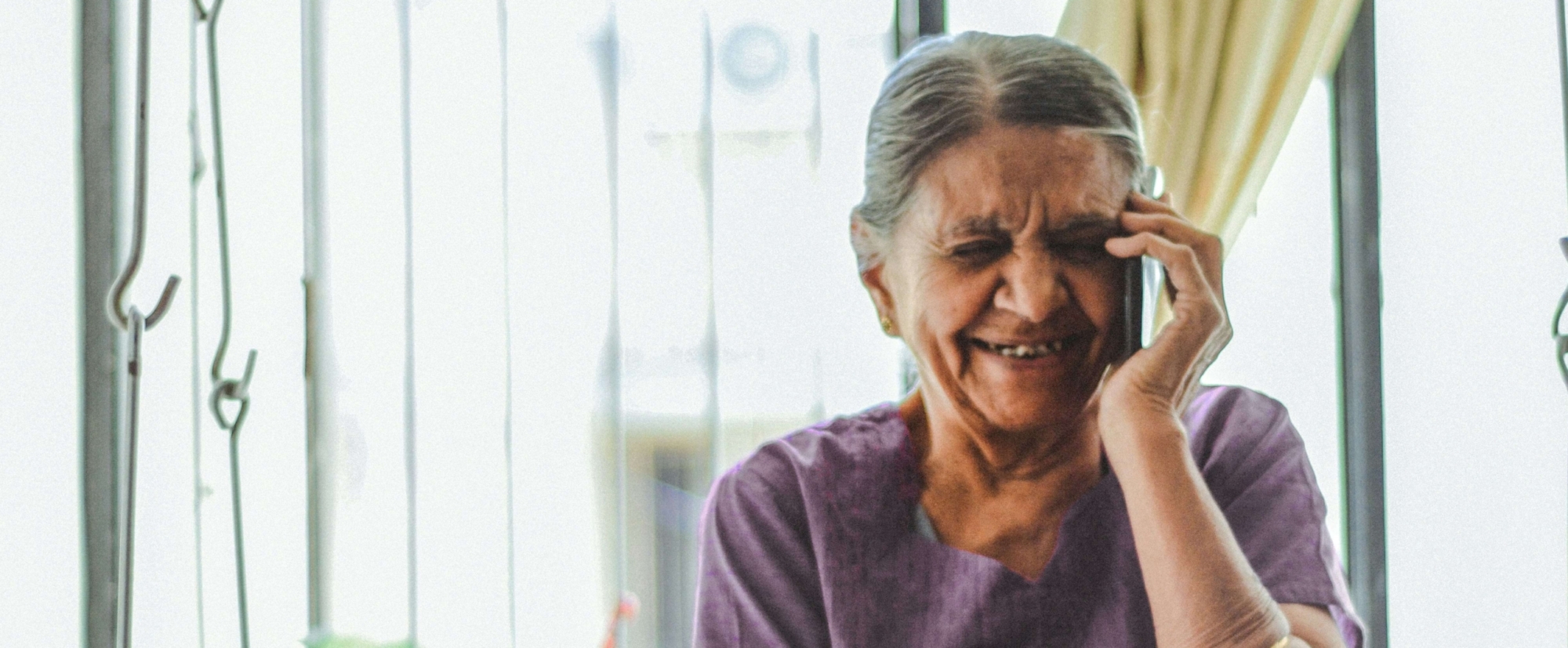 An older Indian woman laughing on the phone while sitting by a window.