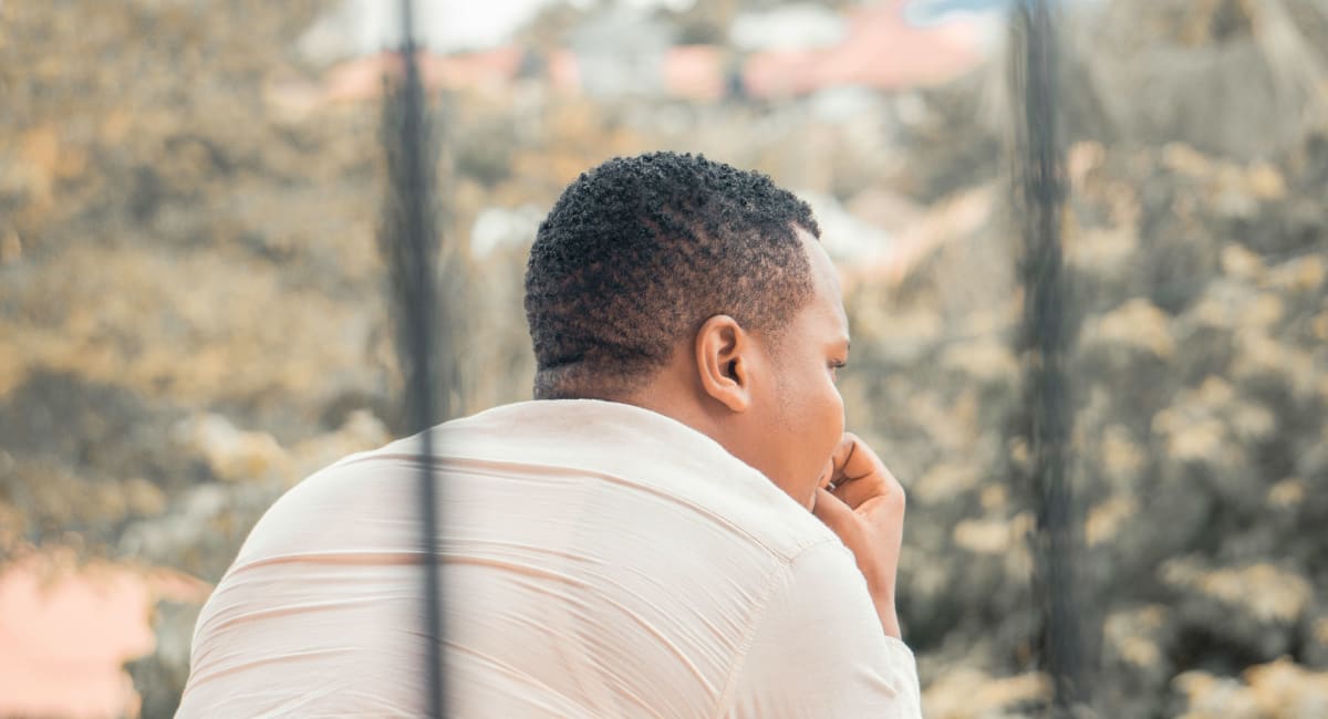 A man with short afro hair facing away from the camera, looking out onto a garden. Most of his face is obscured.
