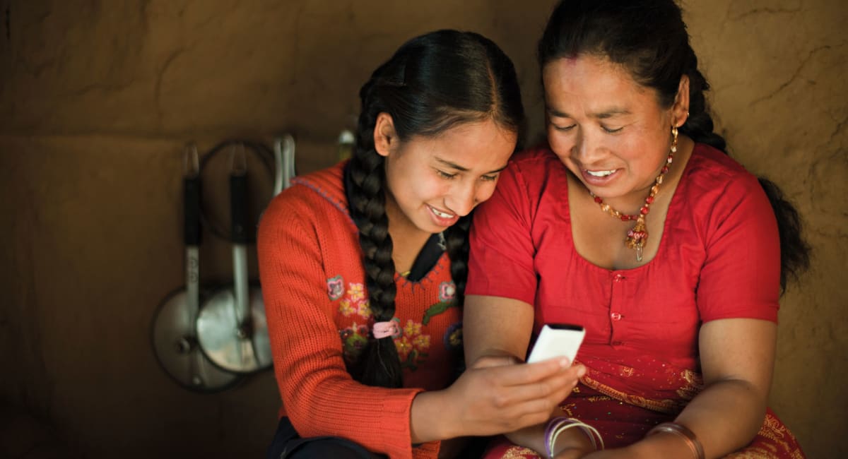 A woman and her daughter smiling as they both look at a mobile phone. They both has dark braided hair and are wearing bright red outfits.