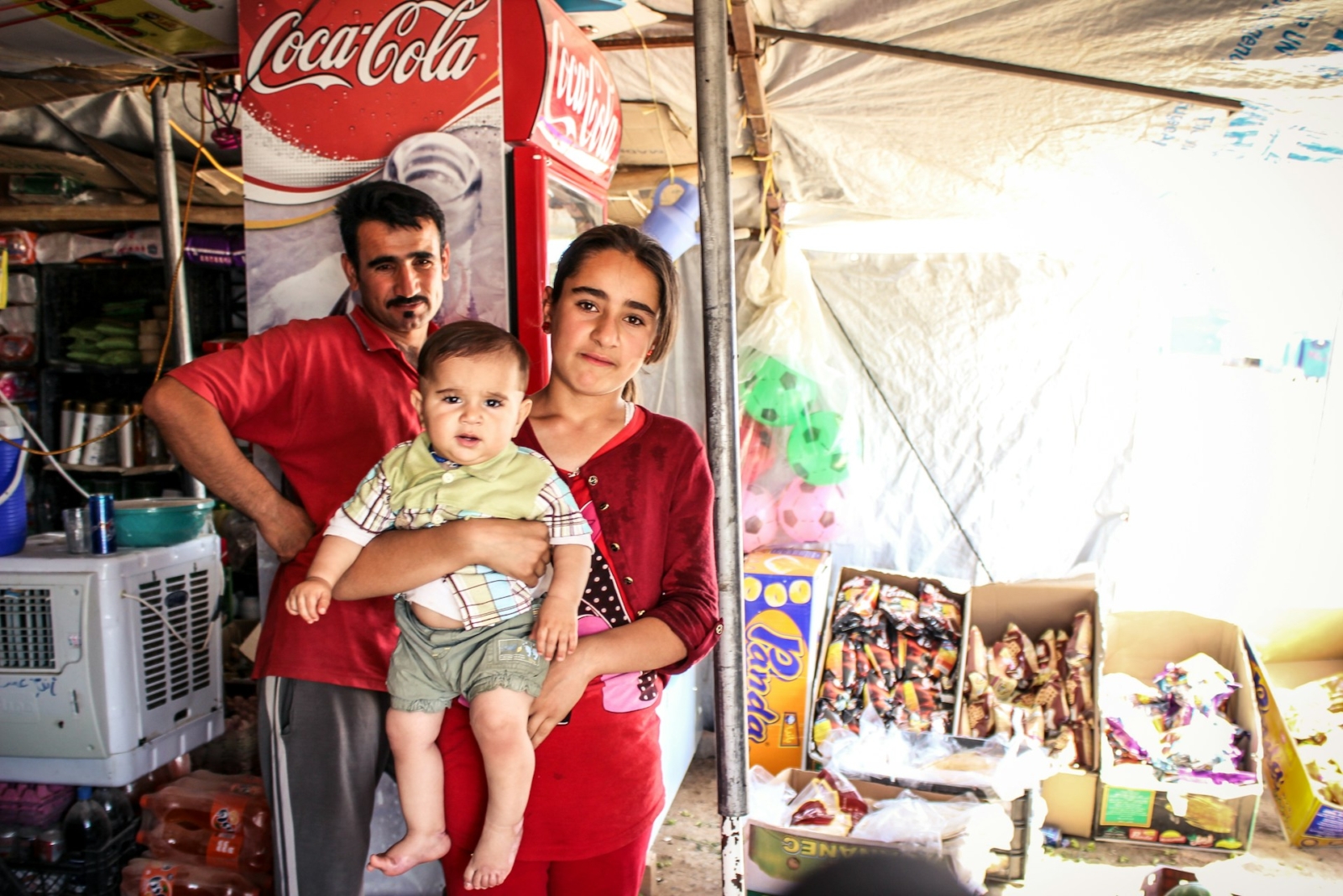 A man and teenage girl holding a baby stand in a small shop next to a Coca-Cola fridge with other snacks and produce in boxes behind them.