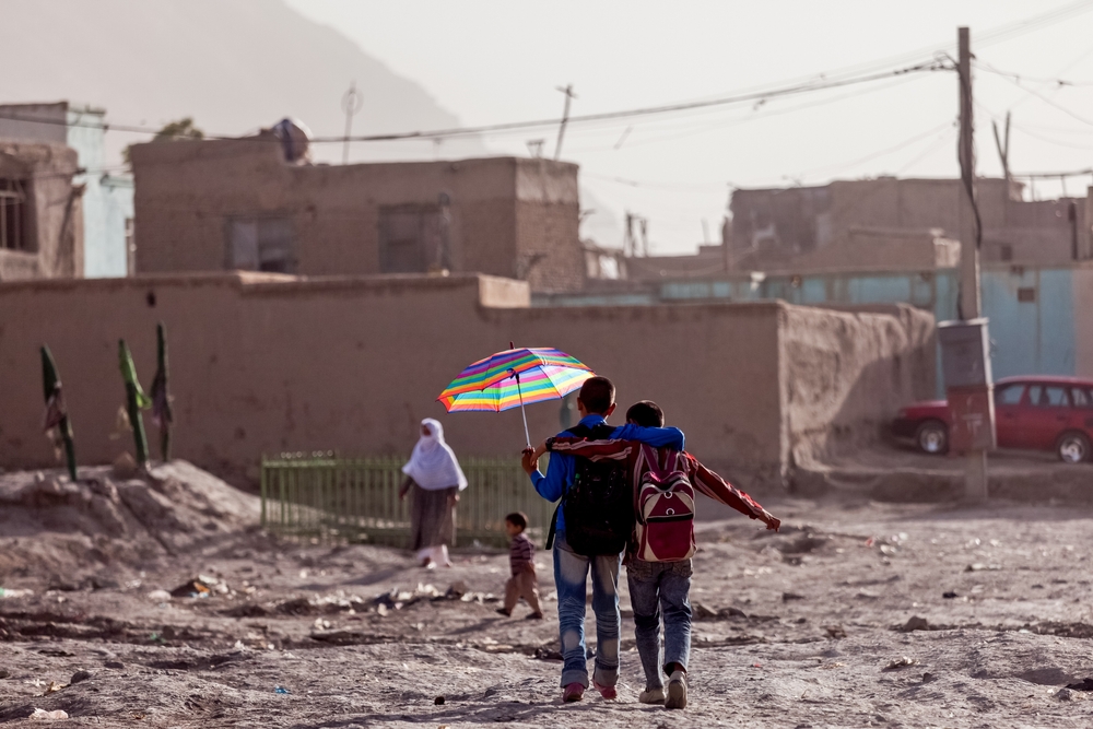 Two boys walk away from the camera with their arms around each other. One is holding a rainbow umbrella to shield himself from the sun. The ground is arid and dusty and there are flat roofed buildings in the distance.