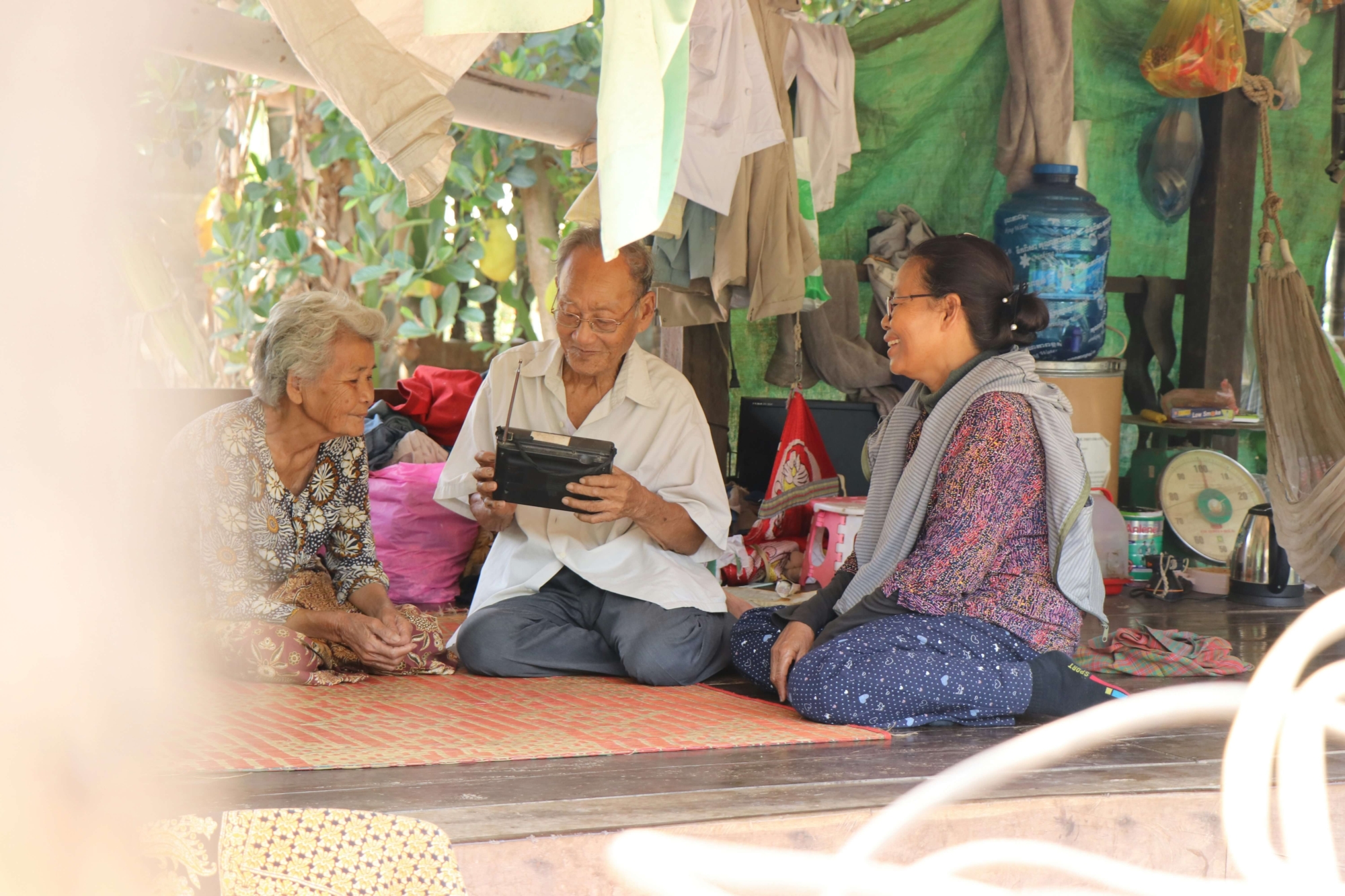 Three people sit on the floor of a traditional house in Cambodia smiling as they listen to an old black radio.