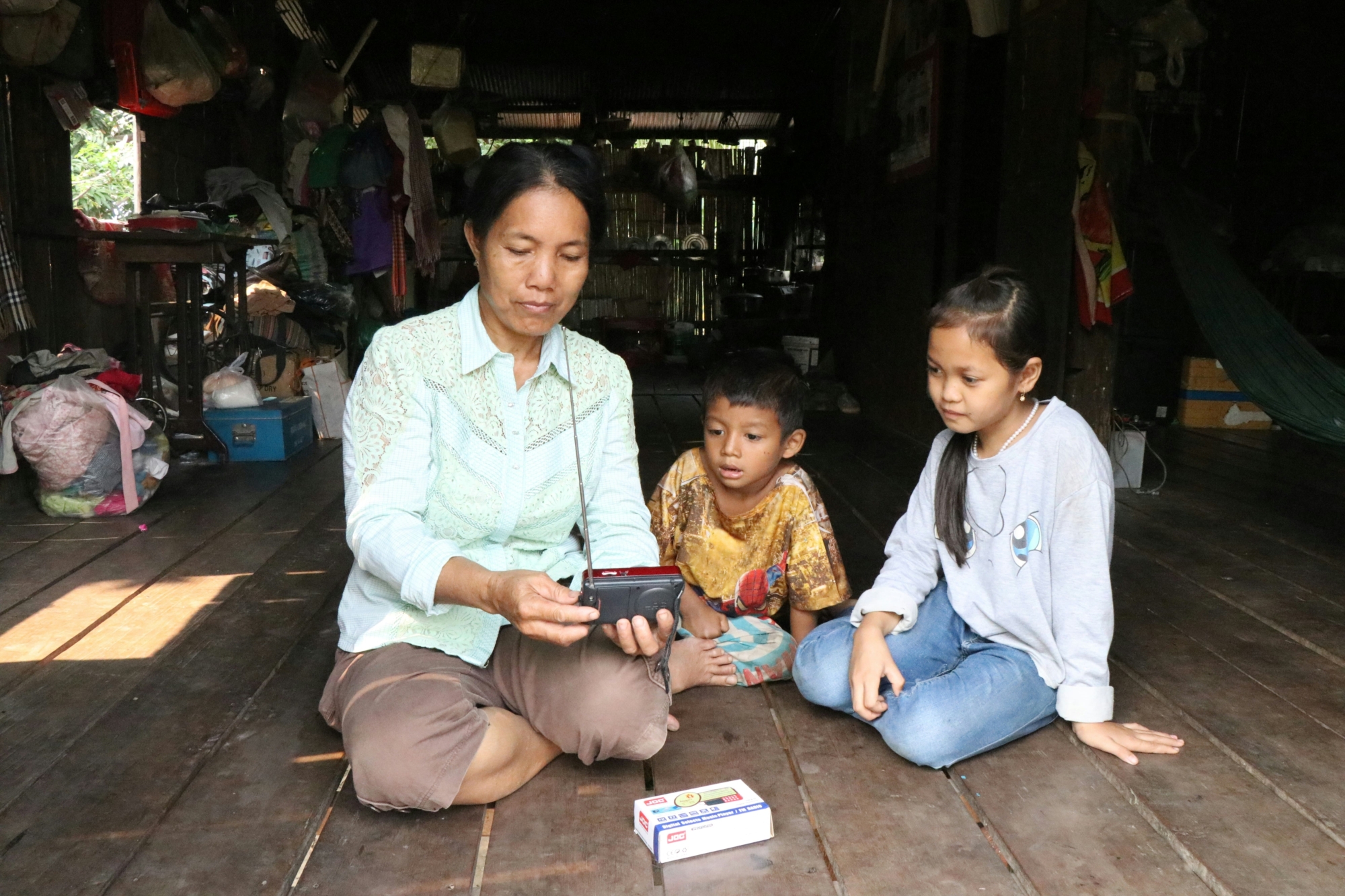 A woman and two children sitting on the floor of a rural wooden house in Cambodia attentively listening to a radio.