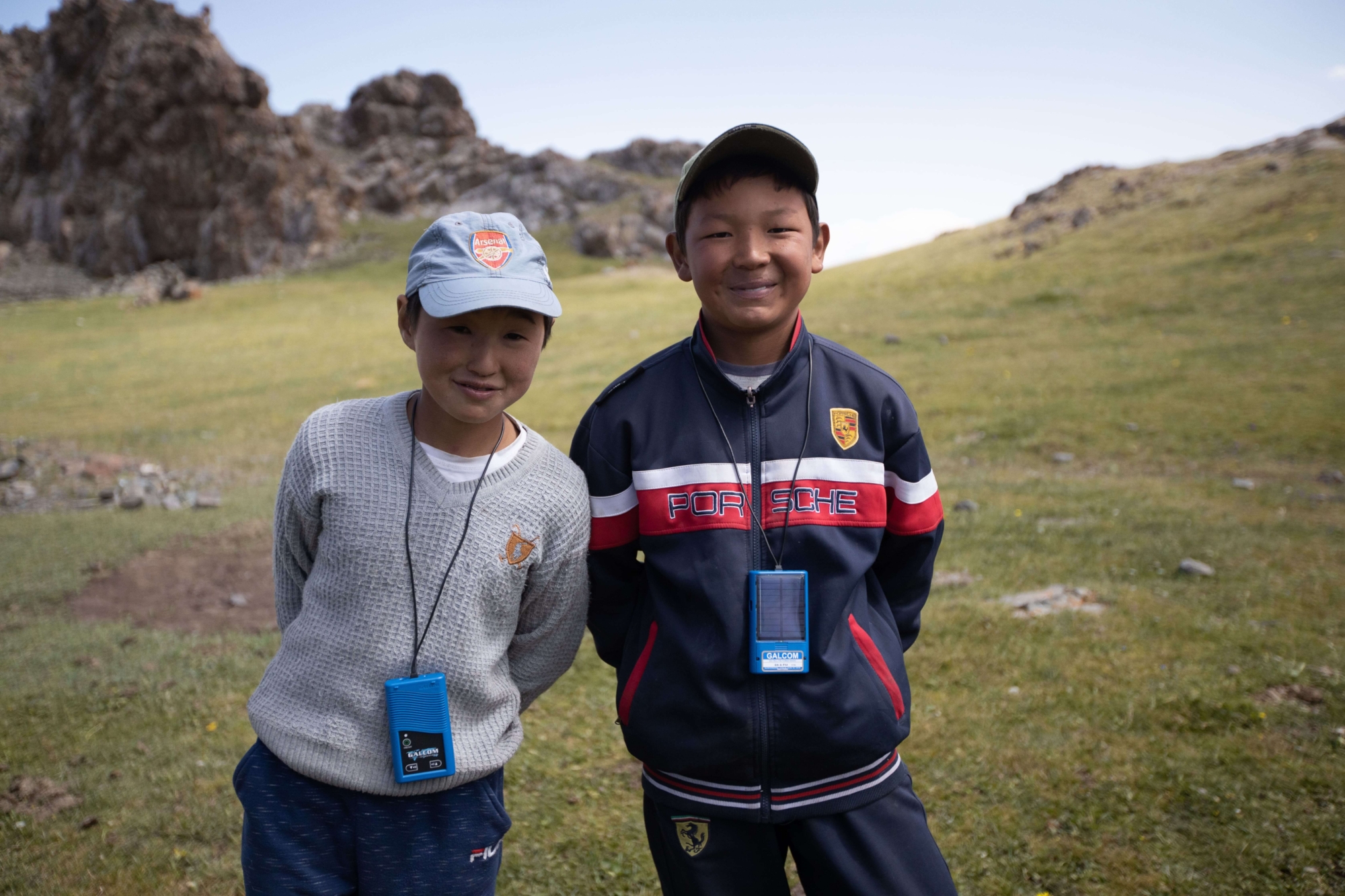 Two young boys on a rocky hill in Kyrgyzstan wearing caps cheekily smile at the camera with their hands behind their backs. They have portable audio devices around their necks.