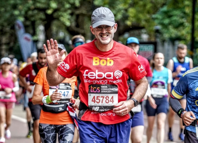 Bob, a man in a grey cap and glasses, smiles and waves while running the London Marathon. He is wearing a red top with the Feba logo printed on it. He is surrounded by other runners in colourful tops.