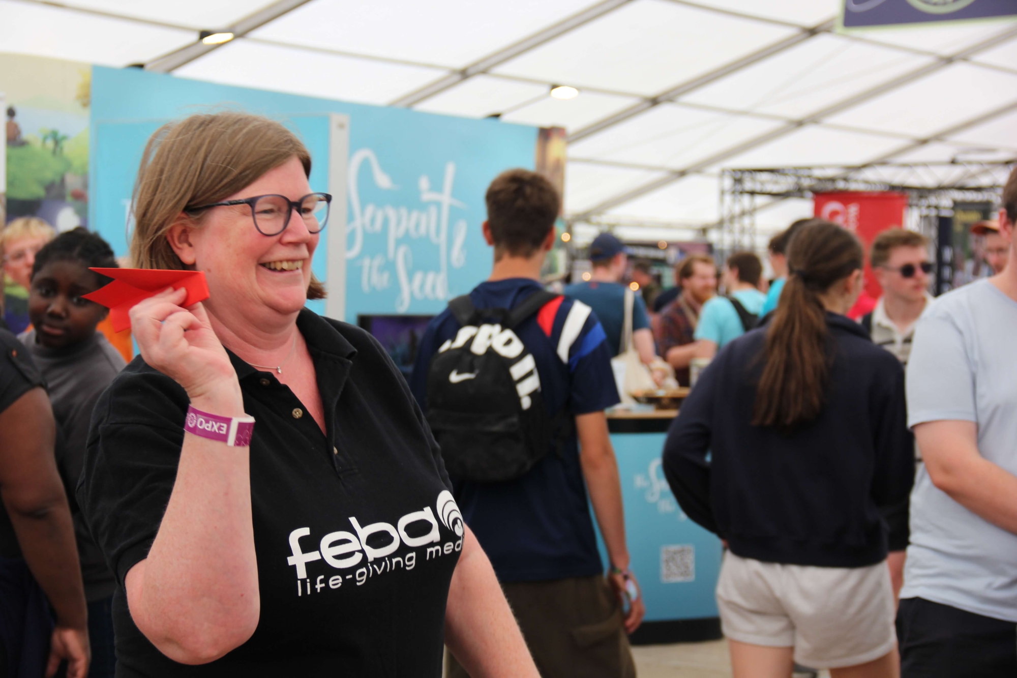 Feba staff member, Catherine, in the exhibition tent at Big Church Festival throwing a paper prayer arrow at the Feba stand. She is smiling and wearing a black polo shirt with the Feba logo on it.