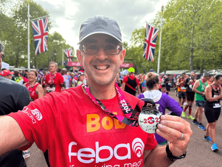 Feba fundraiser Bob taking a selfie at the end of the London Marathon holding his medal. He is standing on The Mall with two union jack flags in the background, surrounded by other finishers.