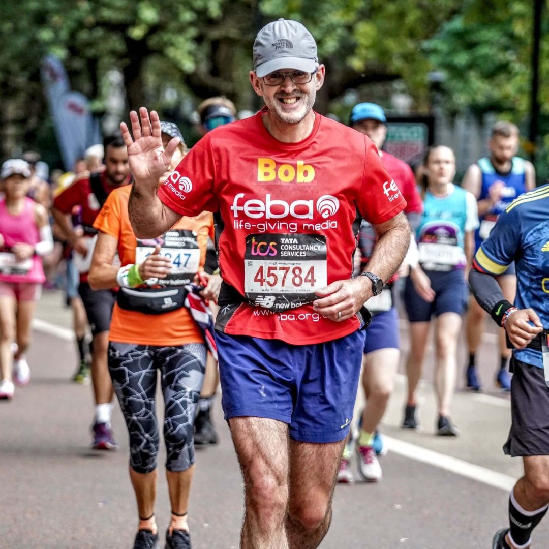 Bob, a man in a grey cap and glasses, smiles and waves while running the London Marathon. He is wearing a red top with the Feba logo printed on it. He is surrounded by other runners in colourful tops.
