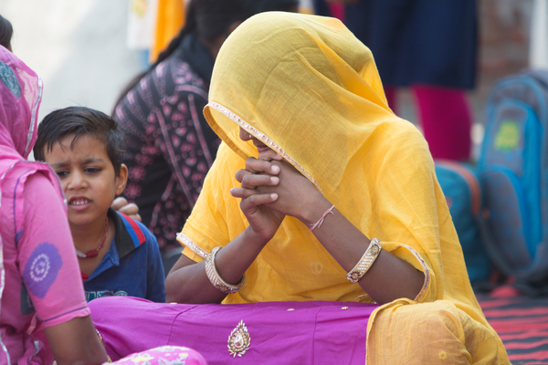 Indian women wearing a bright yellow and pink sari. She is sitting cross legged on the floor with her head bowed and hands clasped under he chin.