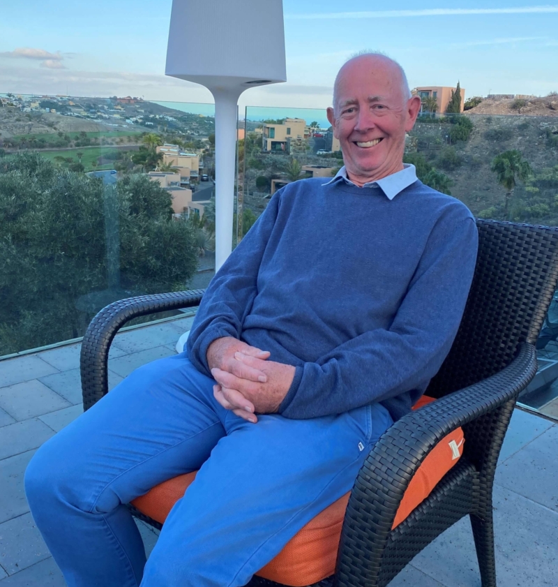 Brian sits in a chair on a balcony, smiling, with a view of mountains in the background. He has his hands folded on his lap, wearing blue chinos, a navy jumper and light blue shirt.