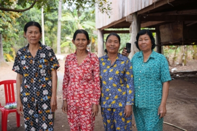 Four Cambodian standing in a line wearing brightly colour matching shirt and trouser outfits. The lady on the end is holding up a portable audio device.