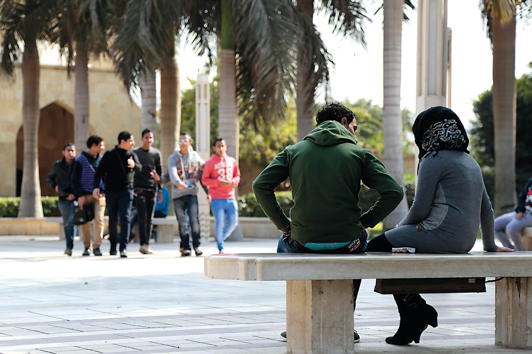 A man and woman sit on a bench facing away from the camera on a paved courtyard in Egypt with palm trees and other people walking in the background.