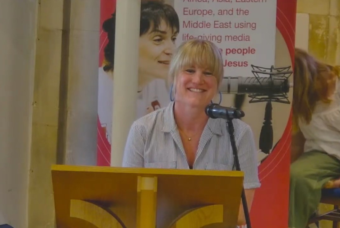 Emily speaking at a church, standing in front of a lectern and microphone with a Feba banner behind her.