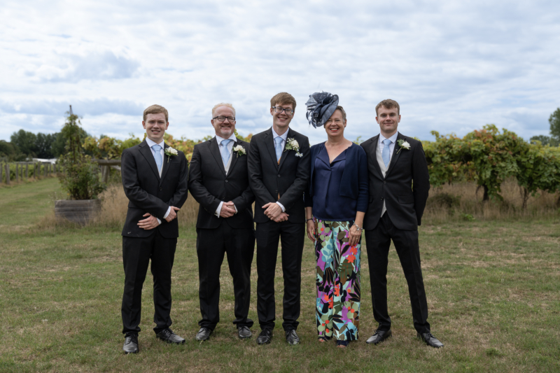 Feba staff member, Esther, with with her three adult sons and husband Paul at her son Ollie's wedding. The men are all wearing black suits. Esther has a navy top and fascinator and colourful floral trousers. They are standing outside in a vineyard.