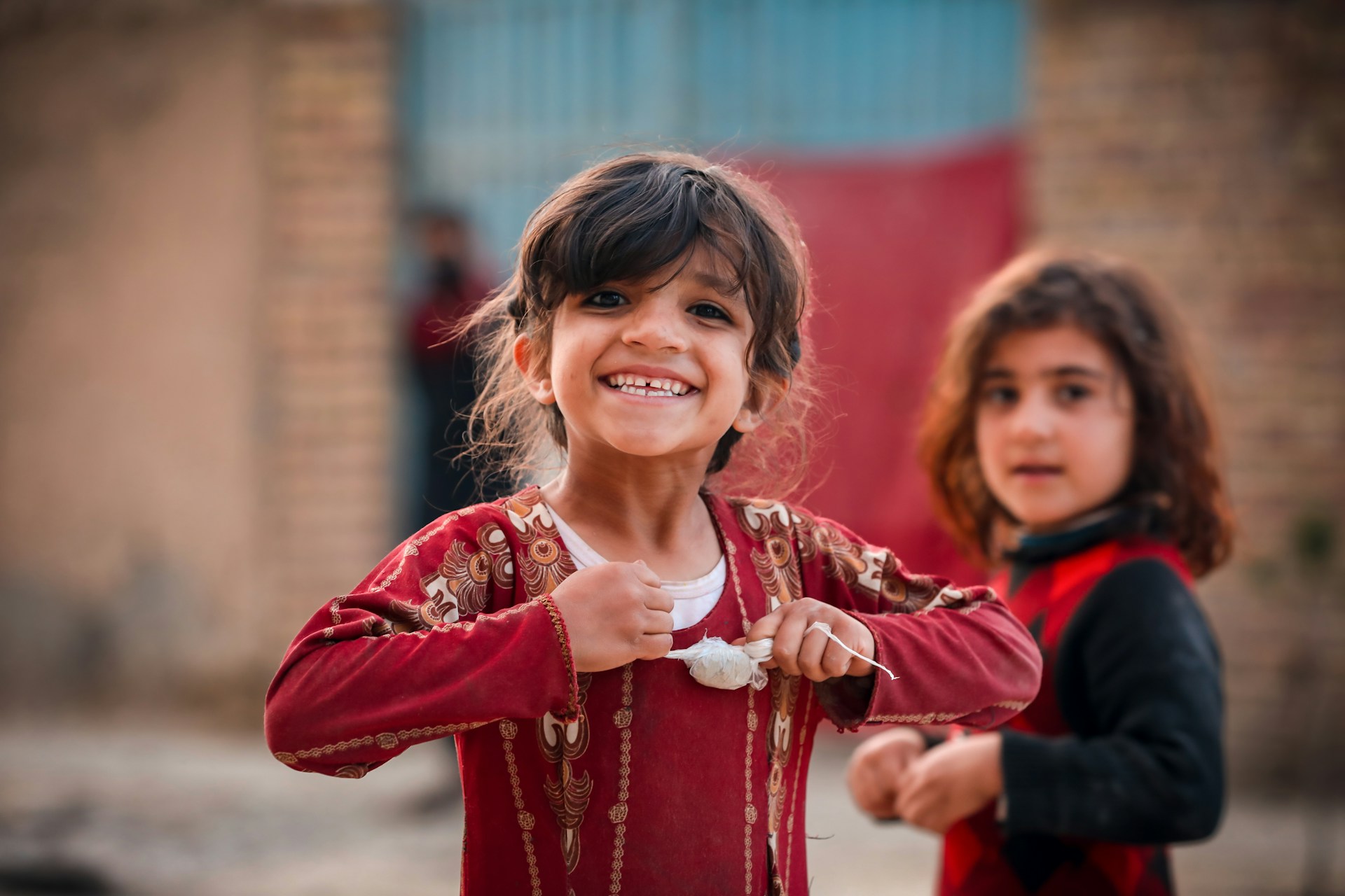 Two Afghan girls playing outside. One grins at the camera while the other stands slightly behind, out of focus.