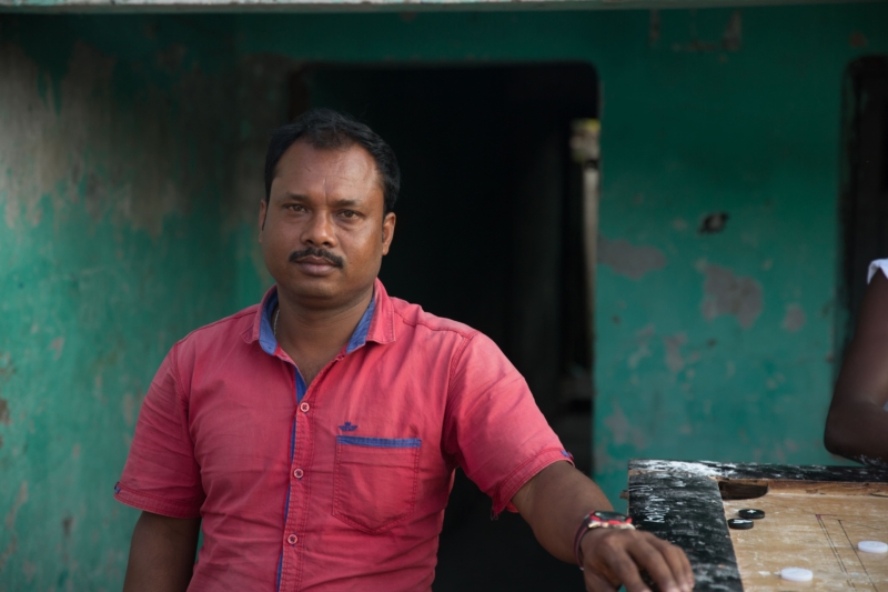 A portrait of an Indian man with dark hair and a moustache, wearing a red shirt, standing outside a teal coloured building.