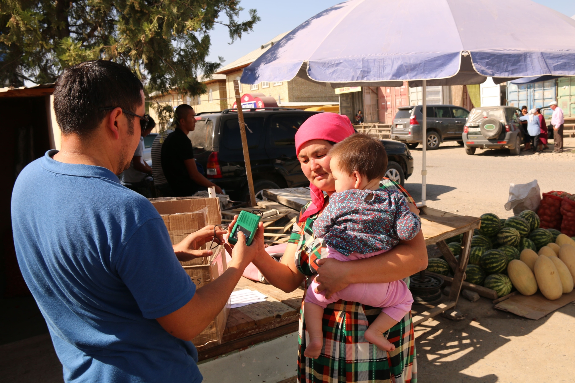 A man in a blue t-shirt hands a green portable audio device to a woman holding a small child. They are standing outside on a street in Kyrgyzstan with cars, market stalls and people in the background.