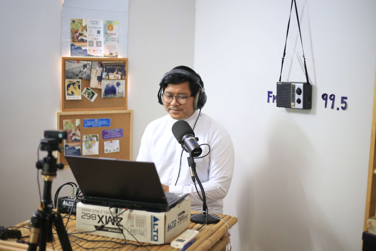Kiry, a member of the Khmer Audio Bible team in Cambodia, sitting at a bamboo desk in front of a laptop recording a radio broadcast. He is wearing a white turtle neck and headphones.