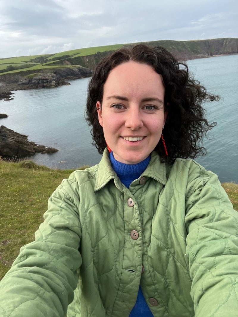 Lucy smiling with a view of cliffs and sea in the background. She has curly brown hair and a green jacket.