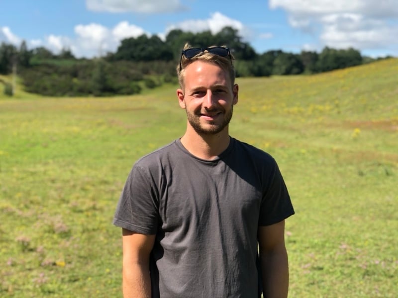 Luke wearing a grey s-shirt with sunglasses on his head smiling in a green sunny field.