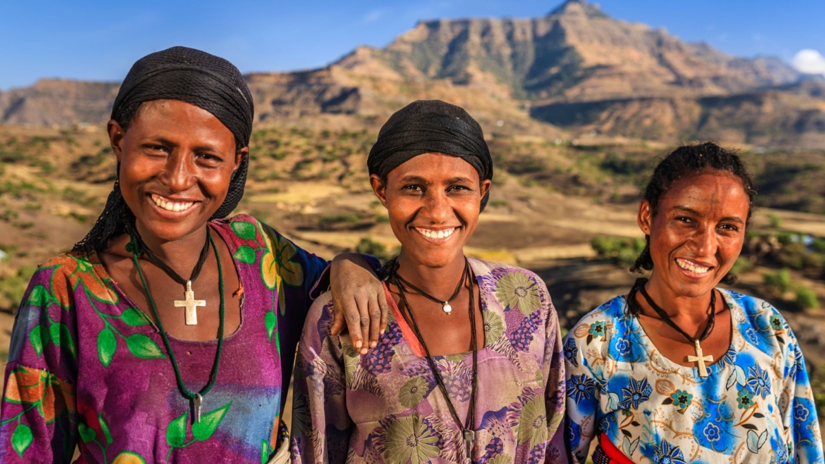 Three Ethiopian women in colourful floral dresses wearing black headscarves smiling with arid rocky mountains in the background.
